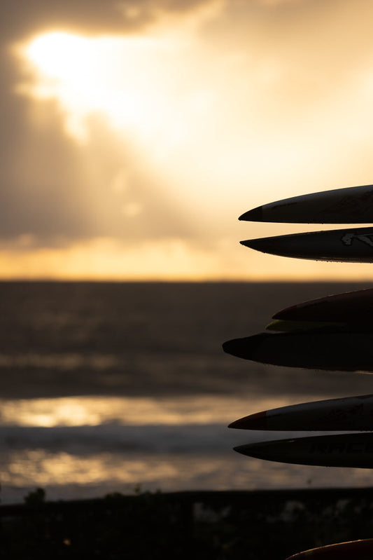 A stack of surfboards in silhouette against a golden sunset sky over the ocean. The tips of the boards are visible, with water droplets on them.