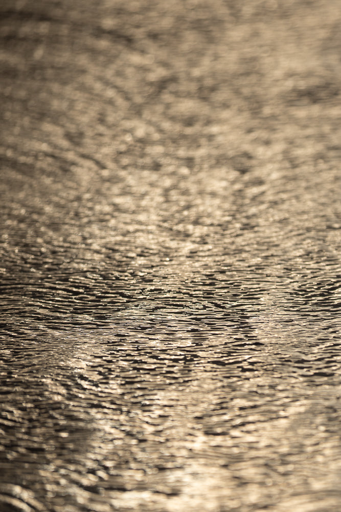 Close-up of rippling water with sunlight reflecting off the surface, creating a textured pattern of light and shadow.