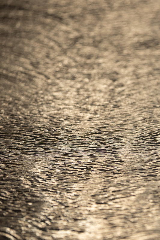Close-up of rippling water with sunlight reflecting off the surface, creating a textured pattern of light and shadow.