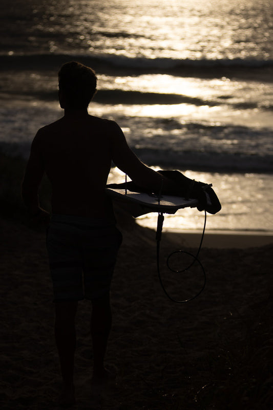 Silhouette of a young man holding a surfboard, walking on a sandy beach towards the ocean at sunset. The sun reflects off the water, creating a shimmering effect.