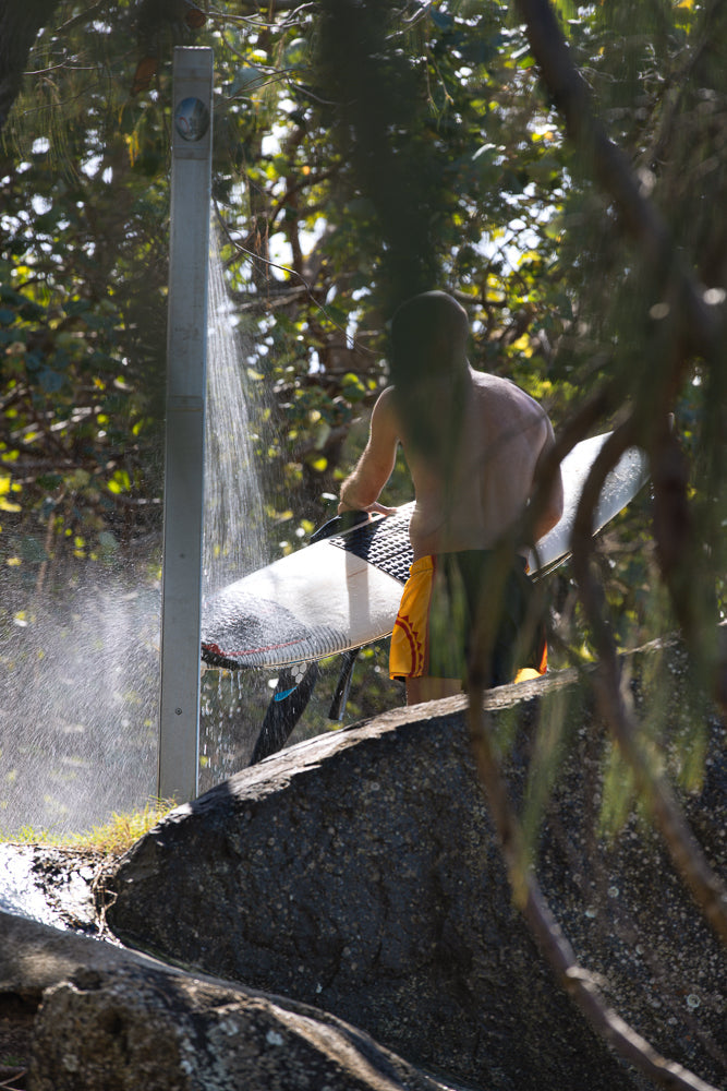 A surfer rinses their surfboard under an outdoor shower, surrounded by lush green foliage and rocks.