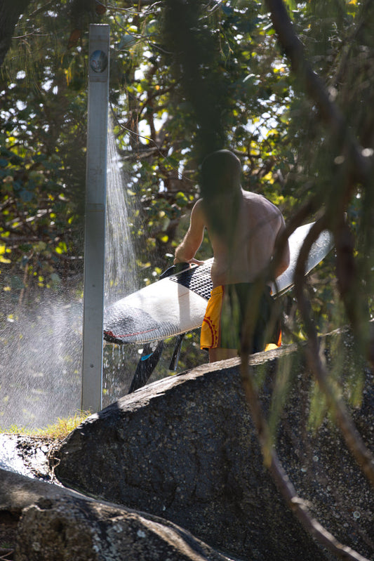 A surfer rinses their surfboard under an outdoor shower, surrounded by lush green foliage and rocks.