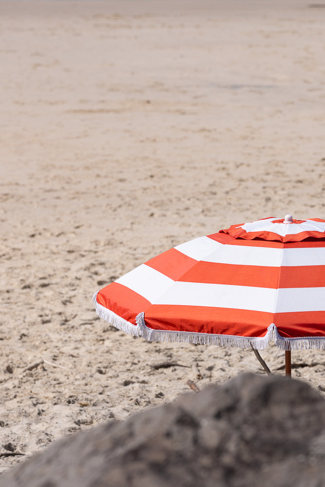 A close-up of a beach umbrella with red and white stripes and white fringe, set against a blurred background of sand.