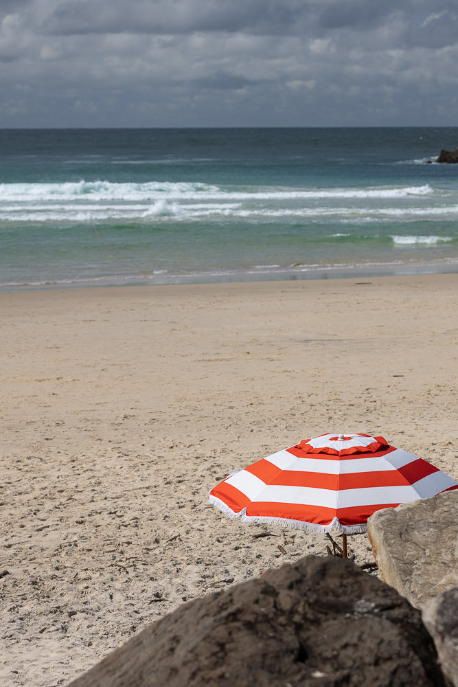 A red and white striped beach umbrella stands on a sandy beach with the ocean and cloudy sky in the background. Rocks are in the foreground.