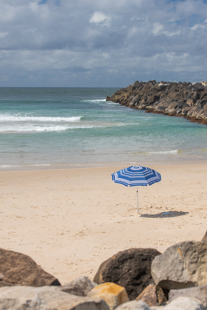 A blue and white striped beach umbrella stands on a sandy beach, with gentle waves rolling in from the turquoise ocean. A rocky breakwater juts out into the sea under a cloudy sky.