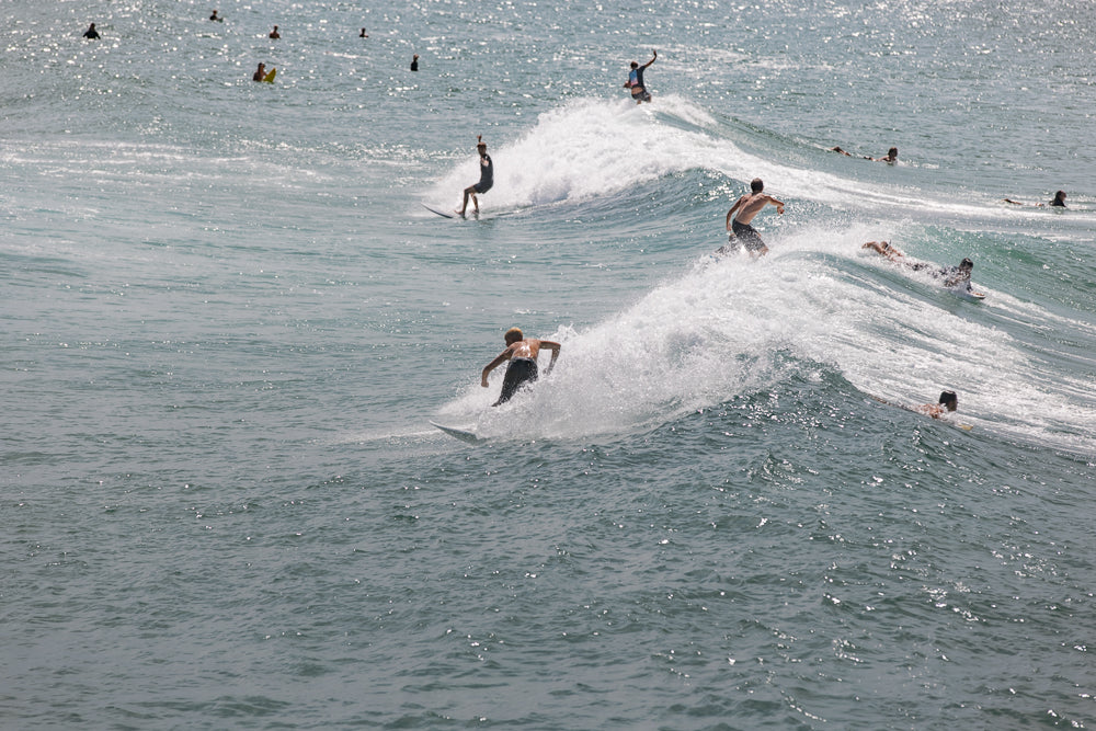 Several surfers ride waves on a sunny day. One surfer in the foreground is captured mid-turn, spraying water as they carve along the face of a wave. Other surfers are visible further out in the ocean, some paddling and others riding smaller waves.