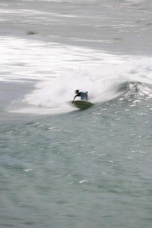 A surfer in blue shorts and a dark top carves a wave, creating a spray of white water. The surfer is positioned on a green surfboard, leaning into the turn as the wave crests.
