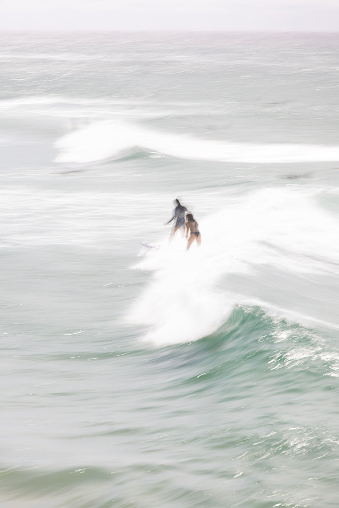 Two surfers, one in a wetsuit and the other in a bikini, ride a wave in the ocean. The image has a soft, blurred effect, emphasizing the movement of the water and the surfers.