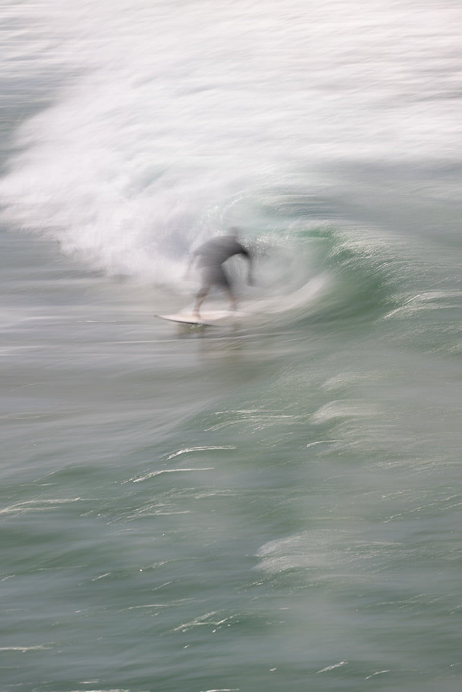 A surfer rides a wave, captured with a motion blur effect that emphasizes the movement and spray of the water. The surfer is silhouetted against the bright white foam of the breaking wave.