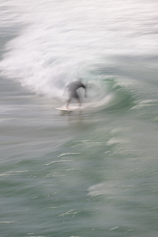 A surfer rides a wave, captured with a motion blur effect that emphasizes the movement and spray of the water. The surfer is silhouetted against the bright white foam of the breaking wave.