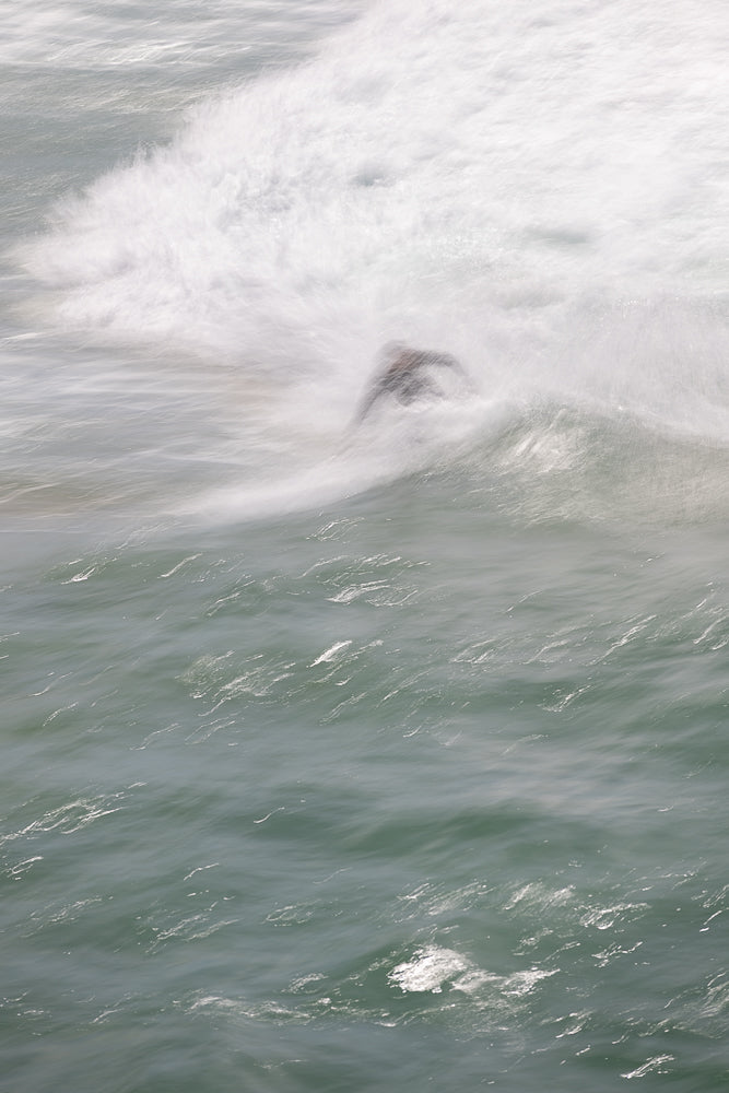 A surfer is captured in motion, blurred by the water's movement as they ride a wave. The image uses a long exposure to create a sense of fluidity and dynamism in the ocean.