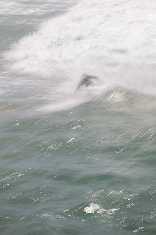 A surfer is captured in motion, blurred by the water's movement as they ride a wave. The image uses a long exposure to create a sense of fluidity and dynamism in the ocean.