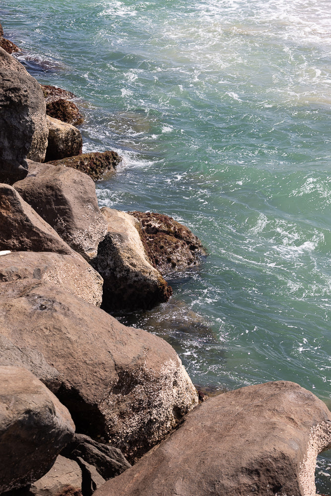 A close-up view of large, weathered rocks covered in barnacles, meeting the turquoise ocean water. Small waves gently lap against the shore, creating white foam and ripples on the surface.