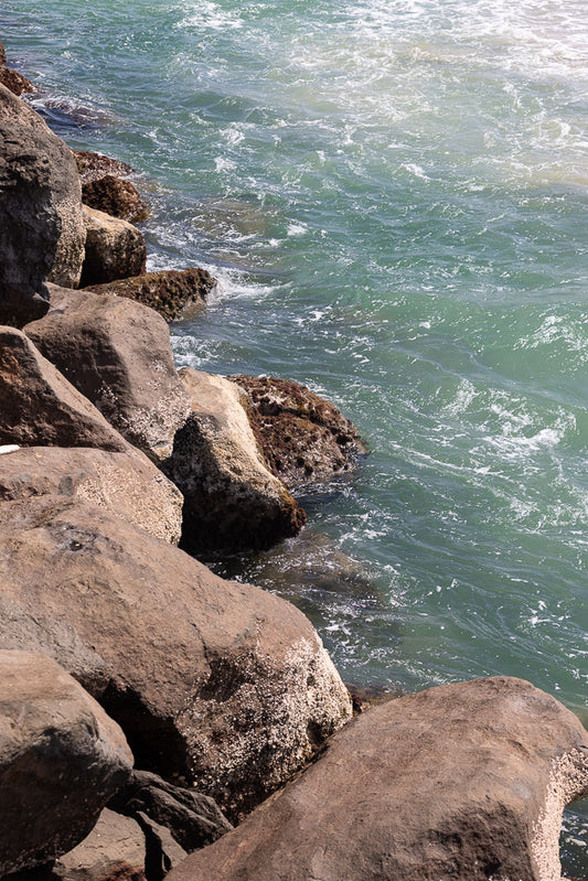 A close-up view of large, weathered rocks covered in barnacles, meeting the turquoise ocean water. Small waves gently lap against the shore, creating white foam and ripples on the surface.