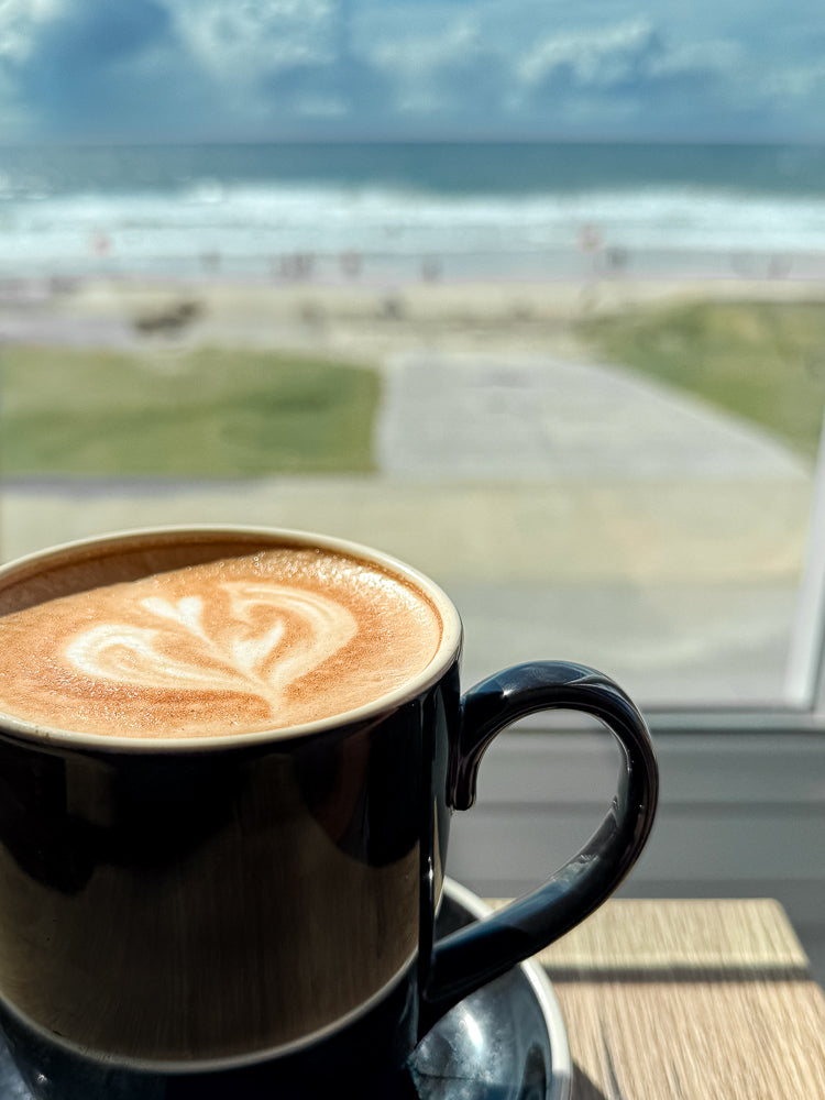 A close-up of a black mug filled with a latte, featuring latte art in the shape of a heart. The mug sits on a saucer on a wooden table, with a blurred background of a beach scene, including the ocean, sand, and people.