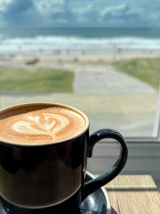 A close-up of a black mug filled with a latte, featuring latte art in the shape of a heart. The mug sits on a saucer on a wooden table, with a blurred background of a beach scene, including the ocean, sand, and people.