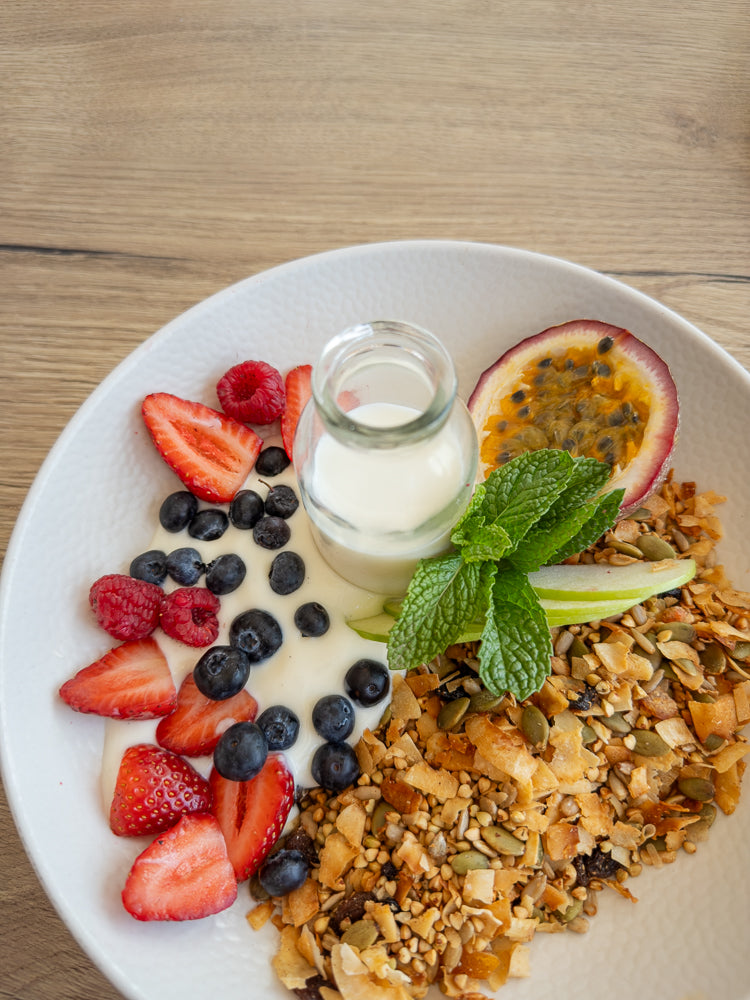 A white bowl filled with granola, yogurt, fresh strawberries, blueberries, raspberries, a slice of passion fruit, mint leaves, and apple slices. A small glass bottle of milk sits in the center.