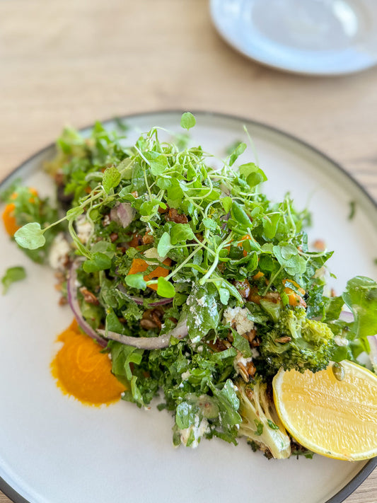A vibrant salad with mixed greens, broccoli florets, crumbled feta cheese, red onion slices, and a dollop of bright orange puree, garnished with a lemon wedge.