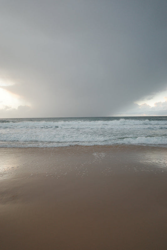 A wide shot of a beach with gentle waves rolling onto the sand under a cloudy sky. The sun is breaking through the clouds on the left side of the frame, casting a soft light on the scene.