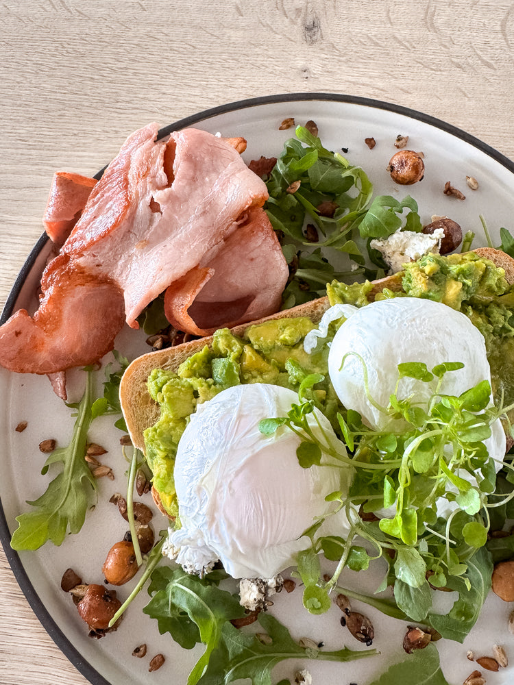 A plate of avocado toast topped with two poached eggs, bacon, and microgreens. The toast is on a light-colored plate with a dark rim, set against a wooden background. Scattered seeds and greens add texture to the dish.