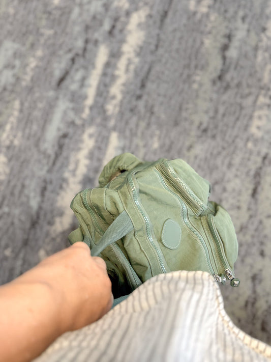 A person's hand holds the strap of a light green backpack. The backpack has multiple zippers and a small circular patch. It rests on a grey carpet.