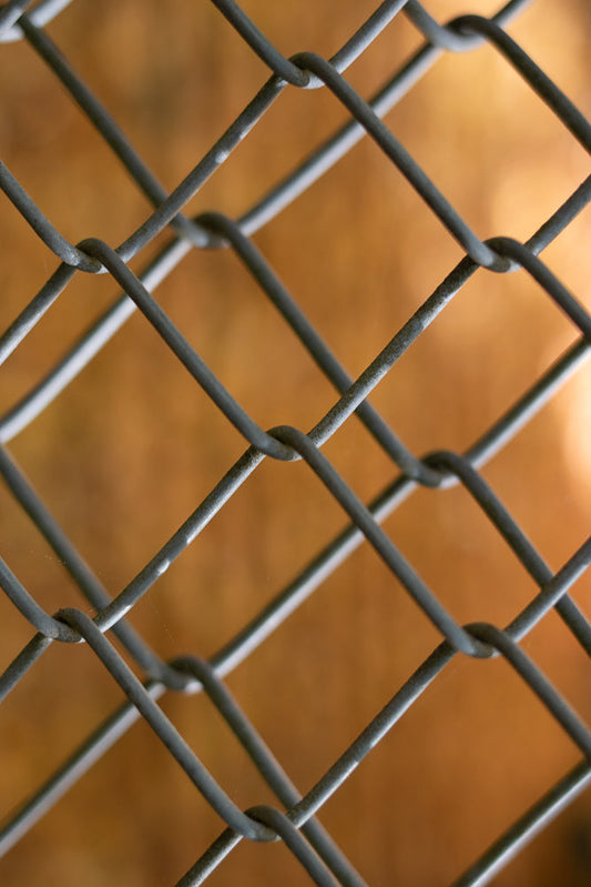 A close-up, abstract shot of a chain-link fence with a blurred orange background. The metallic gray wires form a diamond pattern, with some showing a slight sheen.