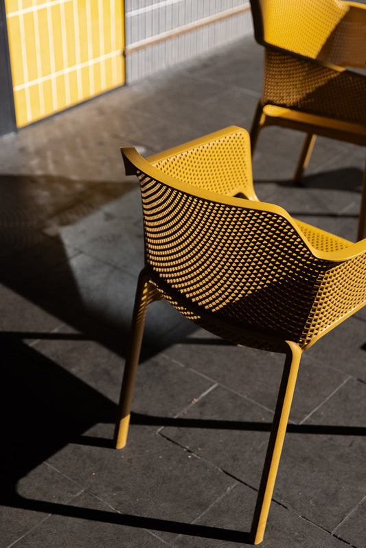 A close-up shot of a bright yellow modern chair with a perforated design. The chair is casting a sharp shadow on the dark gray pavement. In the background, a yellow wall with vertical white stripes is visible.