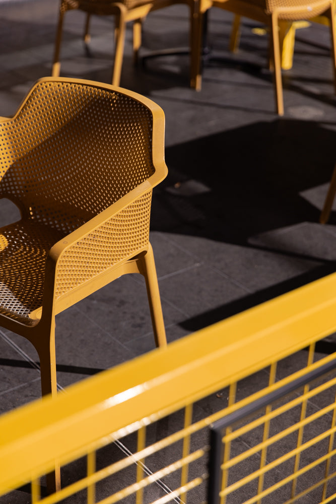 A close-up shot of a yellow perforated chair with a yellow railing in the foreground. The chair is casting a shadow on the dark ground.