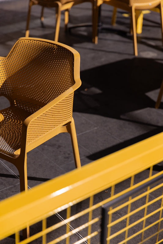 A close-up shot of a yellow perforated chair with a yellow railing in the foreground. The chair is casting a shadow on the dark ground.
