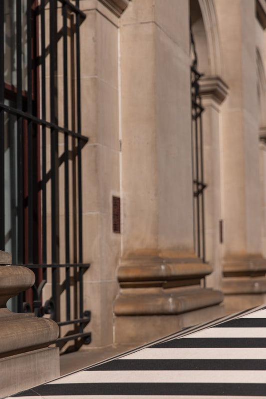 A close-up shot of a building facade with large stone columns and arched windows. A black metal railing is visible in the foreground, casting shadows on the building. The lower part of the image features a black and white striped pattern.