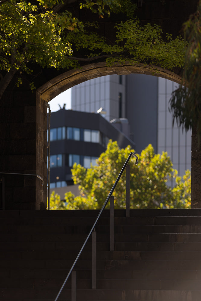 View through an arched stone structure of a metal handrail on dark stairs, with blurred green trees and modern buildings in the background.
