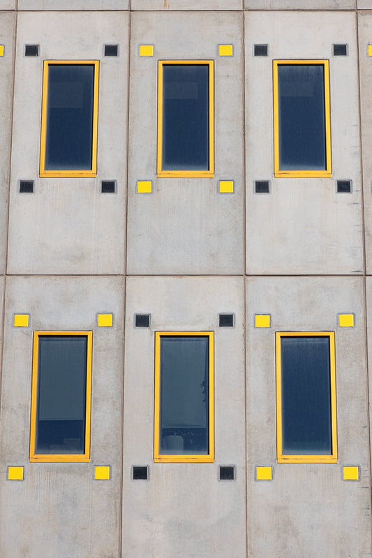 A close-up, straight-on view of a concrete building facade with a repeating pattern of six rectangular windows. The windows have dark blue glass and bright yellow frames. Each window is flanked by two small square accents, alternating between black and yellow.