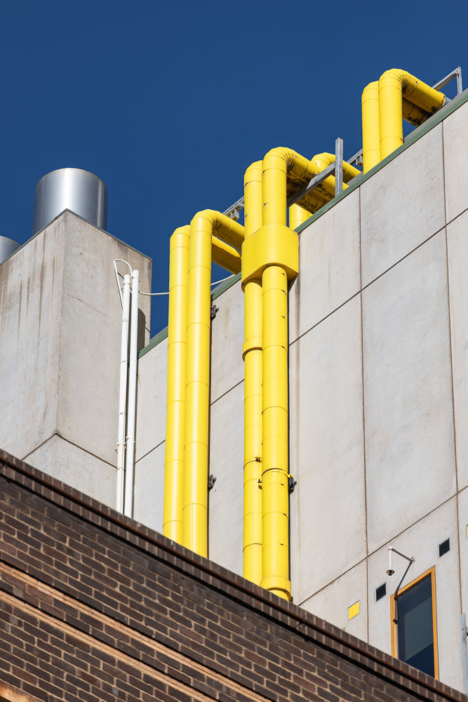 A close-up shot of bright yellow pipes running vertically up the side of a modern building. The pipes are attached to a concrete wall, with a dark brick structure in the foreground. The sky above is a clear, deep blue.