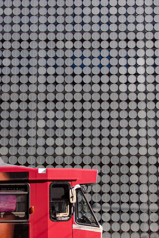 A red bus is parked in front of a building with a facade covered in a grid of circular, metallic panels. The panels have a matte silver finish and are arranged in neat rows and columns, creating a textured, repetitive pattern.