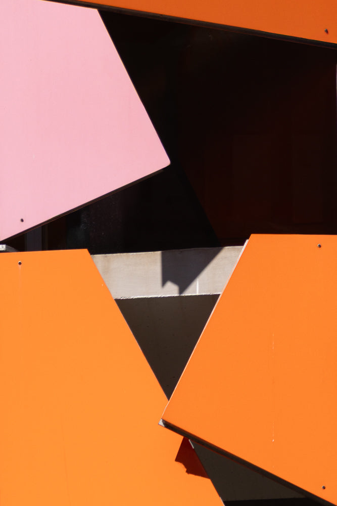 Abstract geometric pattern with large, flat planes of bright orange and pale pink. The shapes overlap and create dark shadows, suggesting an outdoor architectural feature.