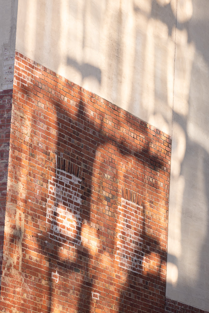 A close-up, angled view of a brick wall with a section of lighter, stucco-like wall. Sunlight casts abstract shadows across both surfaces, creating a textured, graphic pattern.