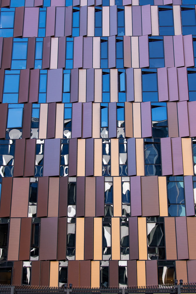 A modern building facade with a repeating pattern of rectangular windows and panels in shades of blue, purple, and brown. Some panels are angled, creating a dynamic, textured appearance.