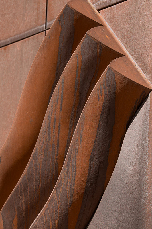 Close-up of weathered Corten steel panels with a textured, rusty surface. Three curved, angular elements with dark streaks of rust are prominent in the foreground, casting shadows.