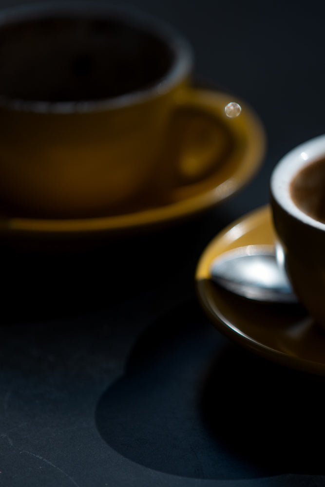 Two yellow cups of coffee sit on matching saucers on a dark surface. A spoon rests on the saucer of the cup in the foreground.