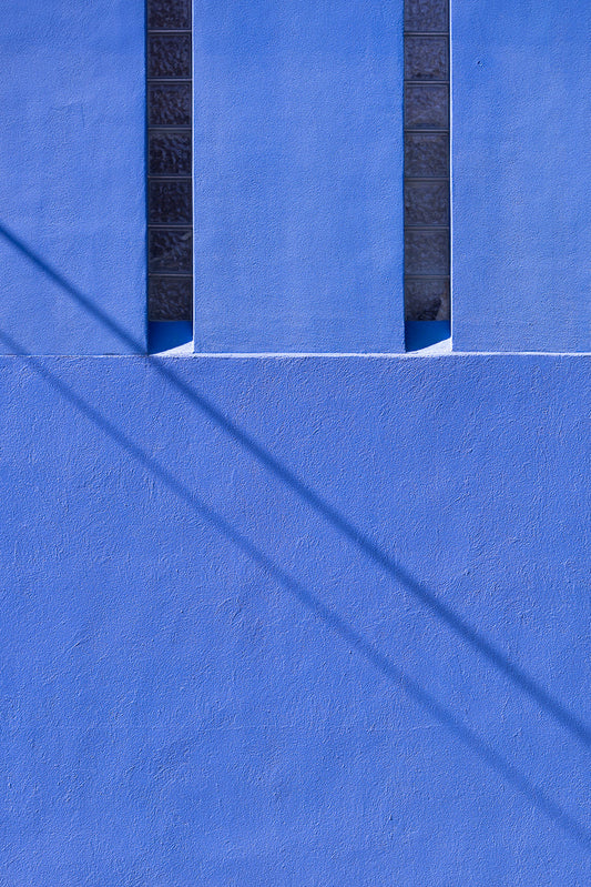 A bright blue textured wall with two vertical columns of glass blocks. A long, dark shadow stretches diagonally across the wall from the upper left to the lower right.