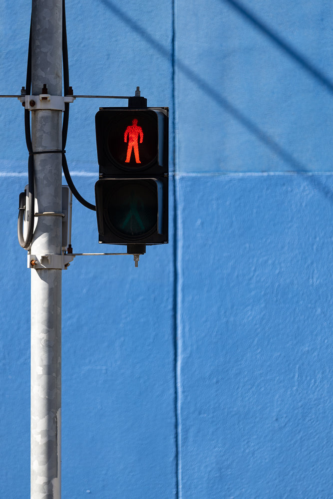 A pedestrian traffic signal displays a red, standing figure, indicating that pedestrians should not cross. The signal is mounted on a metal pole against a bright blue wall.