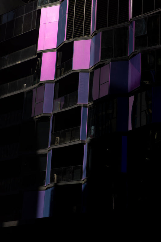 A modern building facade with alternating panels of bright pink and dark blue glass, reflecting light and shadow. The building has multiple balconies with glass railings.