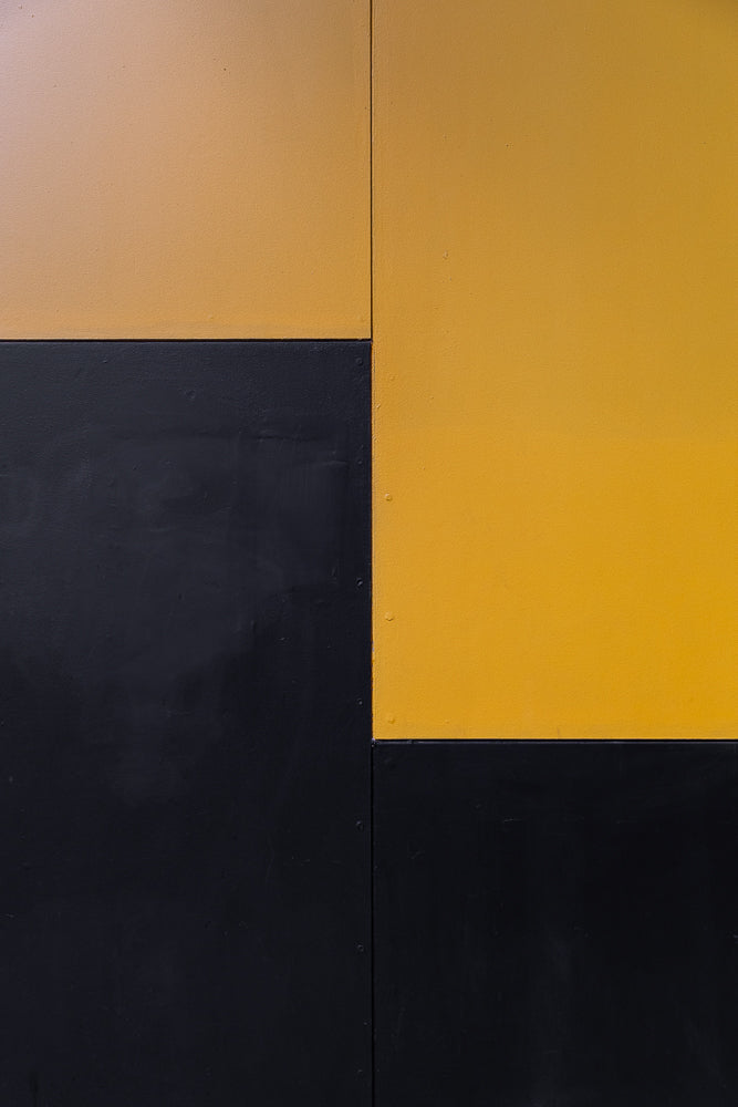 A close-up, abstract shot of a wall divided into four rectangular sections. The top left and bottom right sections are painted a matte black, while the top right and bottom left sections are painted a bright, matte yellow. A vertical seam runs down the center of the image, separating the left and right halves.