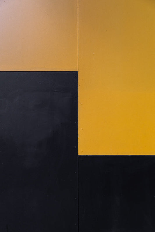 A close-up, abstract shot of a wall divided into four rectangular sections. The top left and bottom right sections are painted a matte black, while the top right and bottom left sections are painted a bright, matte yellow. A vertical seam runs down the center of the image, separating the left and right halves.