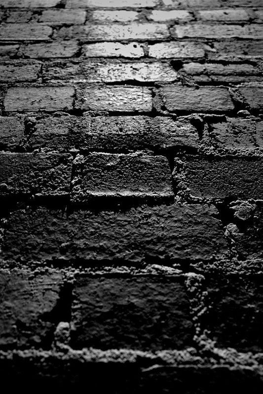 A close-up, black and white, low-angle shot of a rough brick wall. The bricks are dark and textured, with visible mortar lines. A bright light source illuminates the top portion of the wall, creating strong shadows and highlighting the uneven surfaces.