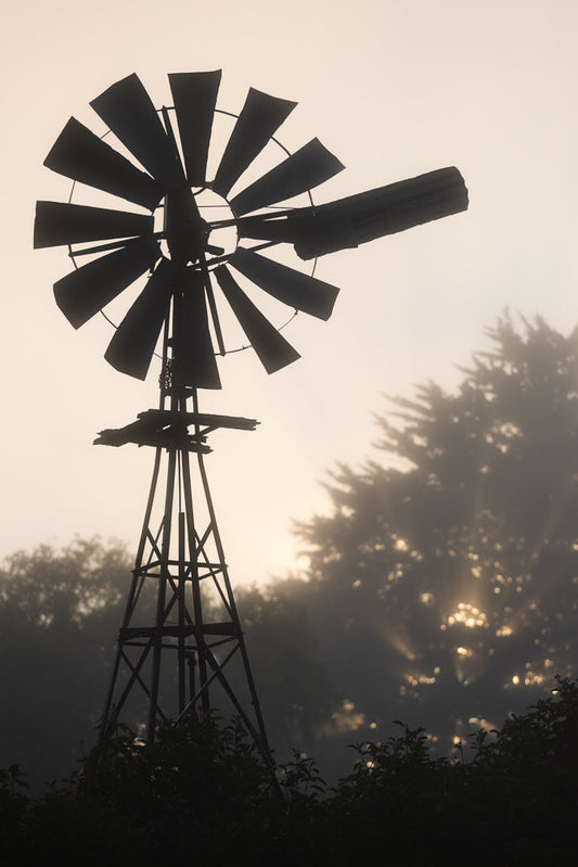 A silhouette of a vintage windmill stands against a hazy, golden sky. The windmill's blades are dark against the soft light, and trees are visible in the misty background, with sunbeams breaking through the fog.