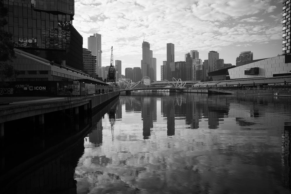 A black and white cityscape shows a river reflecting modern buildings and a bridge under a cloudy sky. A crane stands prominently in the foreground.