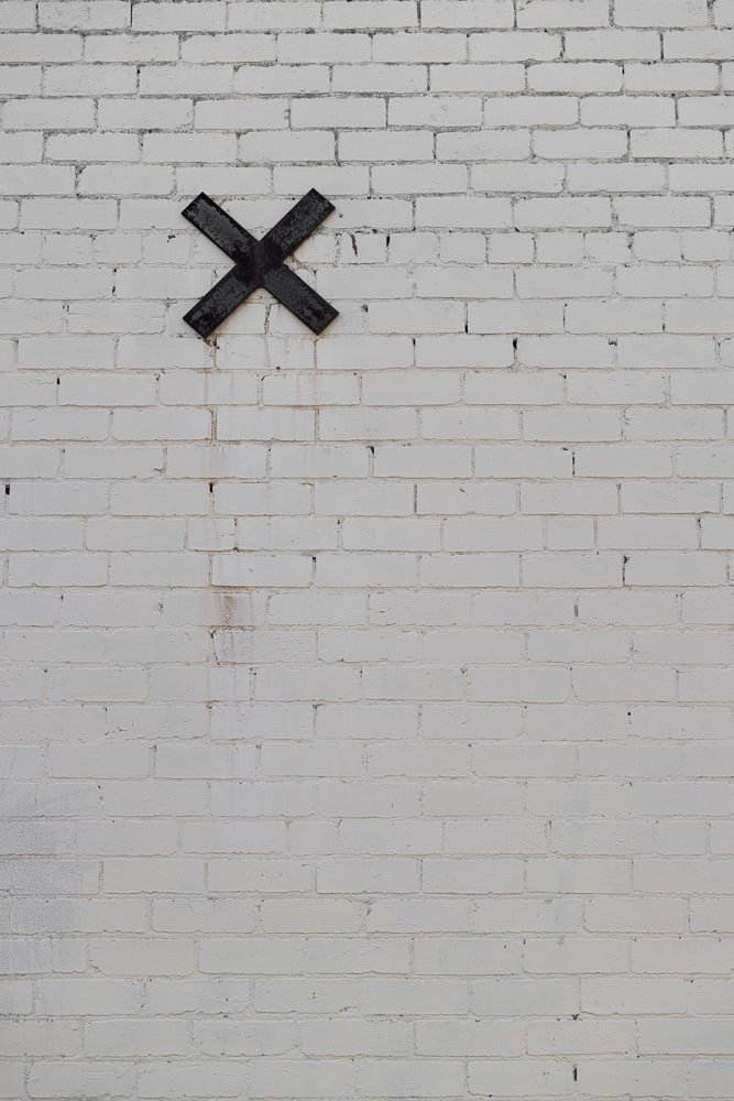 A black metal X is affixed to a white brick wall. The wall has visible mortar lines and some minor staining.