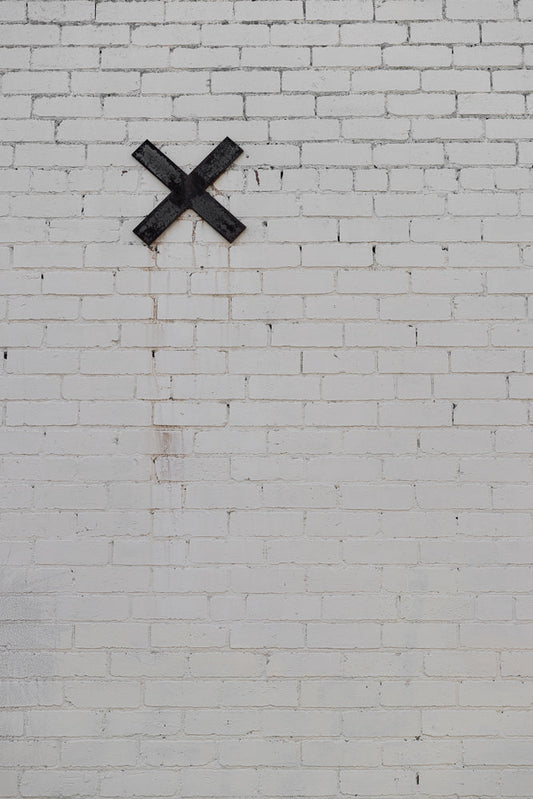 A black metal X is affixed to a white brick wall. The wall has visible mortar lines and some minor staining.