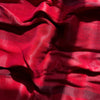 A close-up shot of a red fabric with a textured weave. The fabric is draped and folded, creating shadows and highlights that emphasize its texture and sheen. The lighting suggests it is outdoors or near a window.